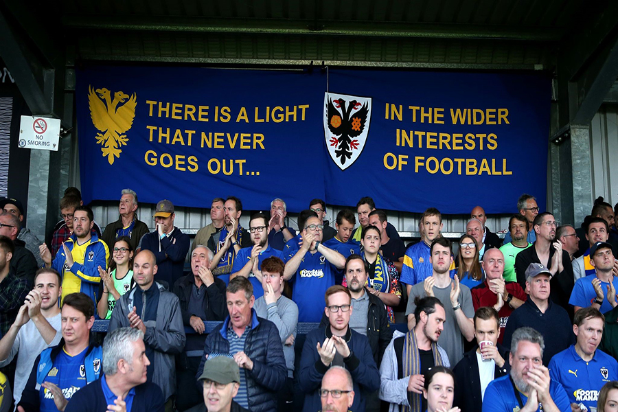 Photograph of football fans at Wimbledon football club with a banner reading "there is a light that never goes out...in the wider interests of football"