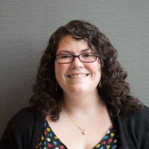 Photo of Carissa Sharp, a white woman with curly shoulder-length brown hair and glasses. She is wearing a colourful blouse, black cardigan, and a small necklace, and is smiling into the camera.
