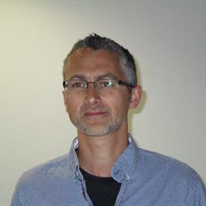 Photo of Jon Catling, a white man with short grey hair and stubble, wearing glasses. Jon is waring a blue shirt and is looking behind the camera
