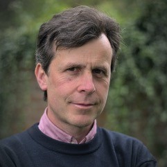 Photo of Miguel Farias, a white man with brown short hair. He is wearing a dark blue jumper with a pink shirt undernearth, and is looking piercingly into the camera.