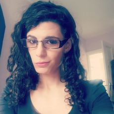 Photo of Sarah Charles, a young white woman with long black curly hair and glasses. She is wearing a black shirt and giving a knowing smile into the camera