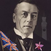 Headshot of a man wearing a monocle with a silhouette of a clock tower behind. In colour a Union Jack flag and a pink orchid in a buttonhole.