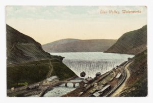 A postcard showing an overflowing dam and reservoir with a bridge and buildings in the foreground