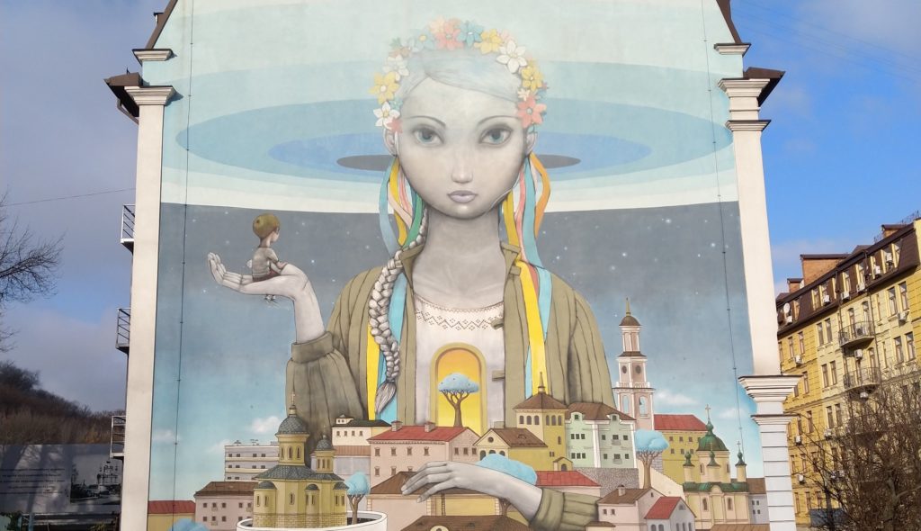 A mural depicting a young woman with blue , yellow, red and white flowers in her hair. A young boy is sat on her open right hand, whilst the other holds a collection of houses.