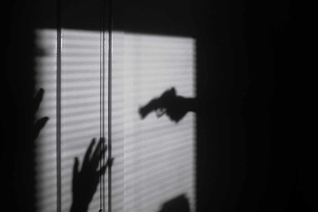 A black and white image showing a shadowed hand holding a gun pointed at another person with their hands raised. 