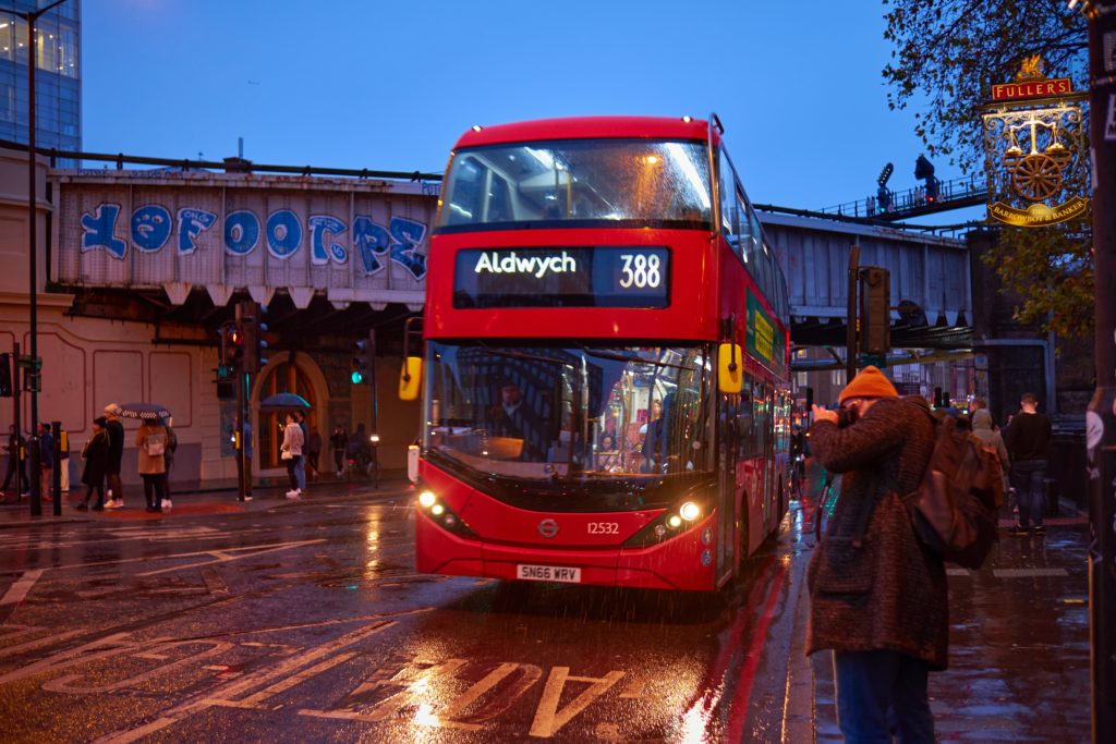 A Red London Bus at a stop in front of a bridge.