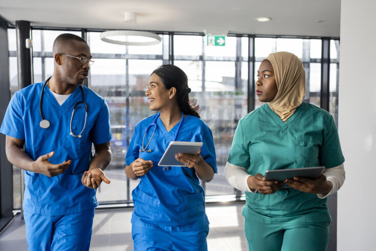 Mixed ethnic group of medical professionals walking down a corridor together in the North East of England. They are working a shift at a hospital and are dressed in scrubs. The women are carrying/using digital tablets.