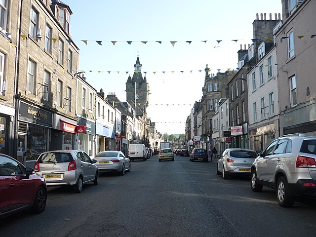 An image of Hawick High Street, cars parked either side of a road in front of shops.