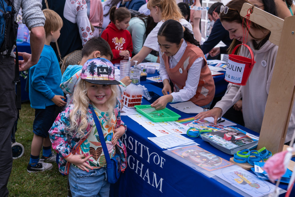 Photo showing several female researchers running family friendly sciences activities for a large group of children who are crowded around the table. Facing the camera is a young girl aged approximately 6 years old who is smiling after having finished taking part in an activity.
