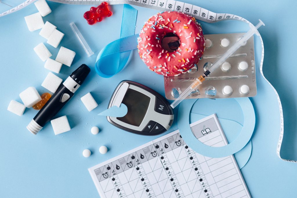 Photograph of different objects associated with diabetes, including sugar, glucose monitor, insulin injection needles, medication and a blue ribbon referencing diabetes awareness