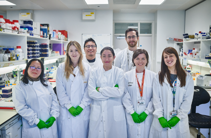 Photo of five female and two male multi-racial scientists in a laboratory setting wearing gloves and lab coats. 