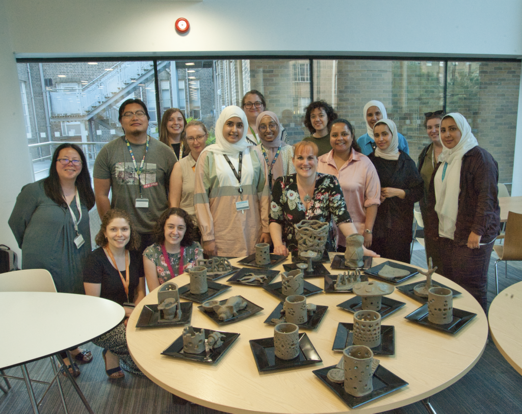 Photograph shows group of 16 people from a wide variety of backgrounds, ethnicities and ages stood together behind a table displaying their clay artworks. The group represents staff and students at the medical school who have taken part in a clay workshop as part of a team building and networking initiative.