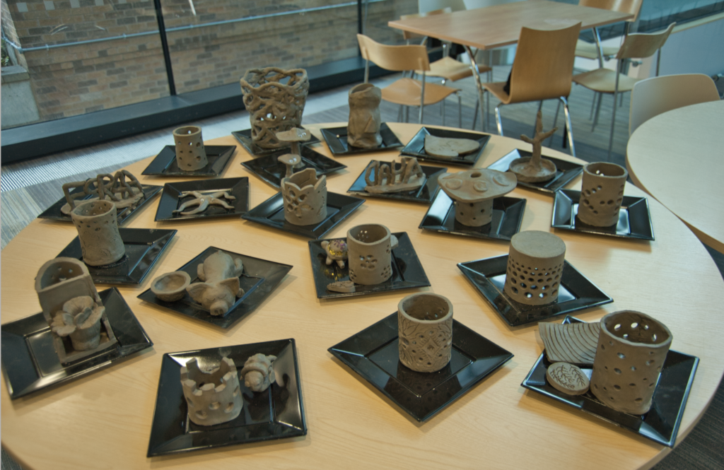 Photo shows a close of approximately 20 different clay artworks ranging in shapes from vases and animal figurines t to miniature castles and other structures. The pieces are presented together on a table and represent the artworks created by hand by students and staff participating in a clay workshop.