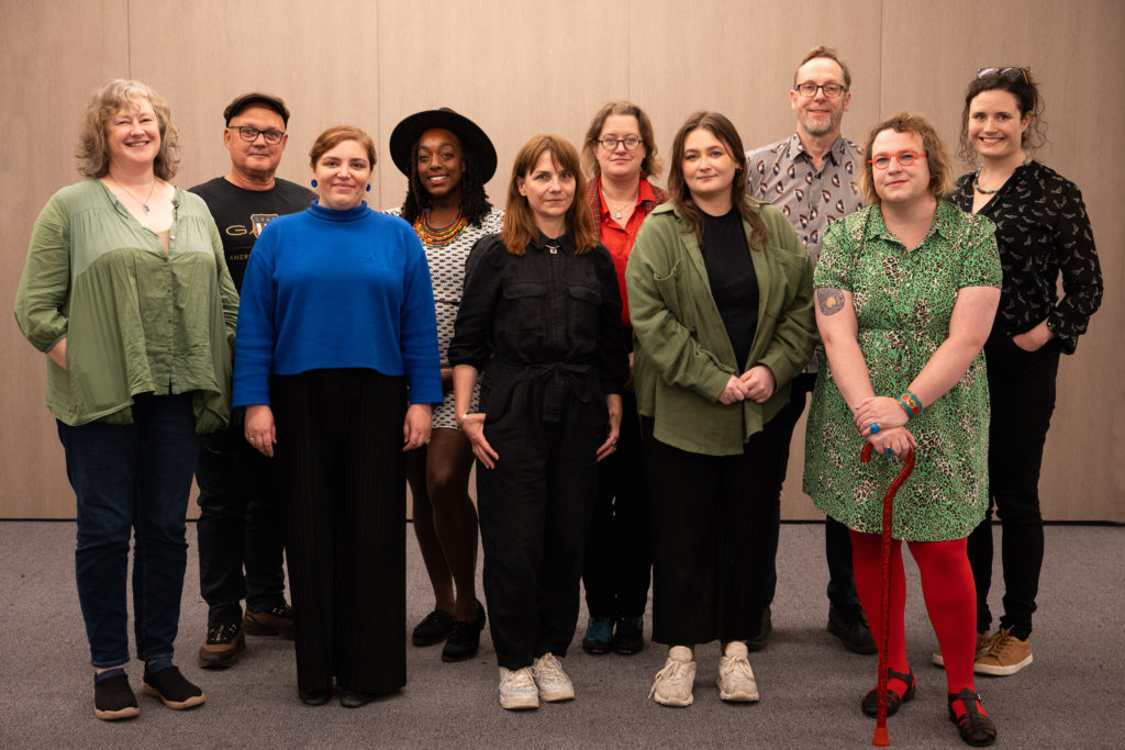 The photo shows the 9 SMQB artist in residence and the programme manager. The artists include 2 males, 6 females and a trans woman. There are also a mix of ethnicities and ages, with one of the female artists being Black and the age range being approximately 25-55 years of age.