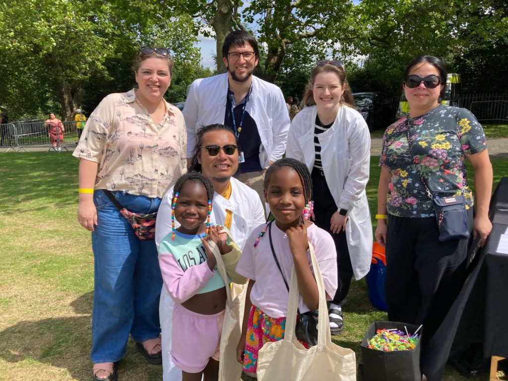 Photograph of 3 female and 2 male IMSR scientists at a festival stall running community engagement activities. Two girls are also in the picture and everyone is smiling.